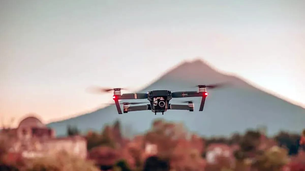 A quadcopter drone with red lights hovering at dusk, with a mountain and buildings blurred in the background