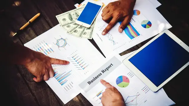 Hands pointing at printed business charts, a phone, a tablet and US dollar notes on a wooden table