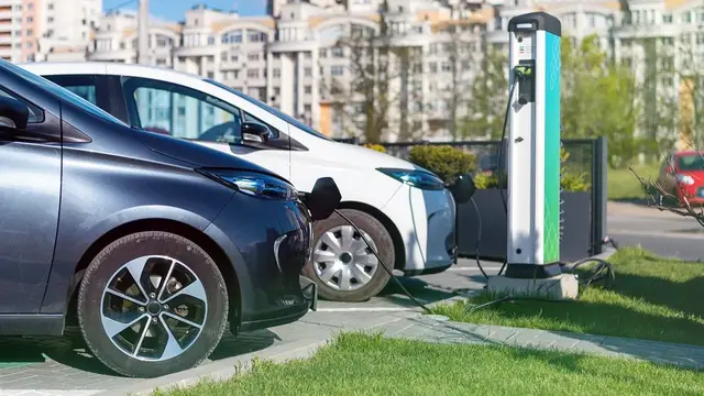 Two compact electric cars plugged into a green-and-white charging post beside a lawn, with apartment blocks behind