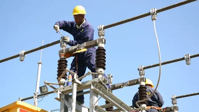Two power-company technicians in hard hats and blue overalls working on high-voltage switchgear against a clear sky