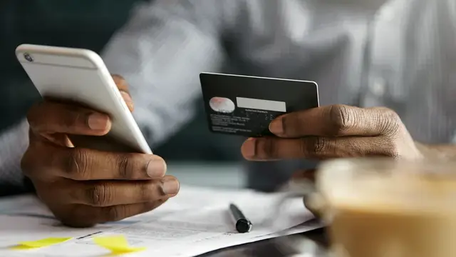 Close-up of a person holding a smartphone in one hand and a bank card in the other, above paperwork and a coffee cup